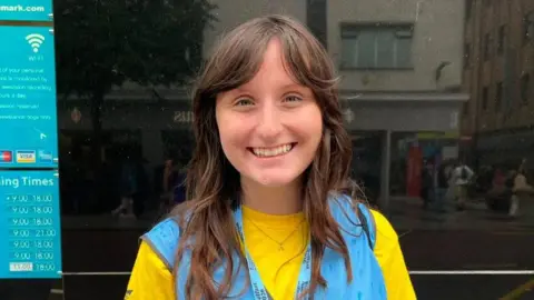 Volunteer now A young woman, Addison McGowan pictured outside Primark home on Royal Avenue. She is wearing a yellow t-shirt with a blue bib on top and a blue lanyard. She has brunette hair.
