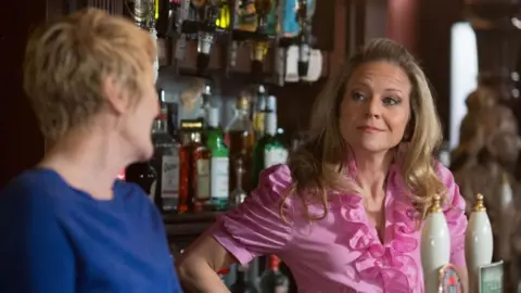 A blonde woman in pink ruffled shirt stands behind the bar of a pub and looks at another woman with her eyebrows raised. 