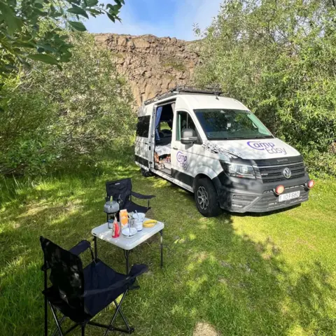 Samantha Phillips-Brown White camper van displaying parked on grass in the foreground, with the side door open. In front of the van, two folding camping chairs and a small table hold a kettle, bowls, cups and food items. Trees surround the campsite, and a rocky cliff rises behind the van. A roof rack is mounted on top of the vehicle.