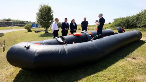 Reuters Wide shot showing Andy Burnham with his Minister for Border Security and Asylum Anna Turley and three officials standing by a recovered small boat. One official is standing inside the boat a black inflatable dinghy that is a few metres long.