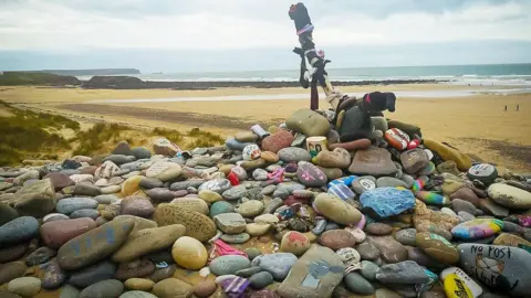 BBC A pile of rocks and pebbles with messages scrawled in colourful writing on them on a beach. A wooden cross sits at the top of the pile.