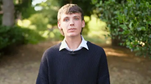 A young man with blond hair and wearing a dark jumper stands on a path in a forest