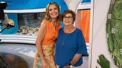 NBC via Getty Images Savannah Guthrie, in a bright orange sleeveless top and patterened orange hued skirt, smiles beside her mother Nancy, in dark-rimmed glasses and a deep blue T-shirt, on a TV news set