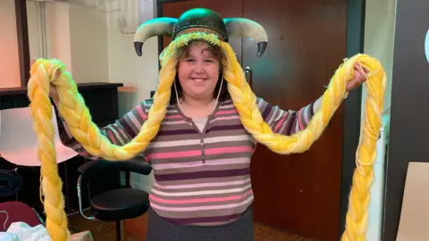 Alex Pope/BBC A child, wearing a striped top, holding out a headpiece with horns and long plaits. She is smiling and looking straight at the camera.