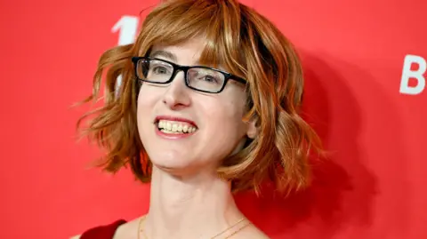 Getty Images A woman wearing a red dress standing in front of a red backdrop. She is wearing a necklace, glasses and has short wavy hair