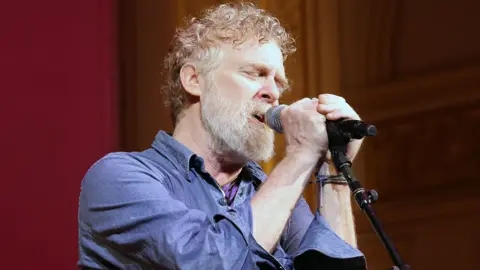 Getty Images Glen Hansard has short curly reddish/brown/grey hair and a greyr beard and is singing with two hands on the microphone and his eyes closed