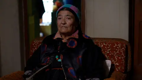 Morup's wife Yangskit dressed in a black Sulma, a traditional Ladakhi robe and a colourful knitted headcover with traditional jewellery around her neck symbolising Ladakh's heritage and Buddhist spirituality