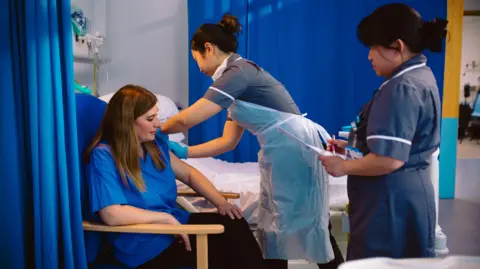 NIHR This image shows a healthcare setting with a seated adult receiving an injection in the upper arm from a healthcare worker wearing gloves and a disposable protective apron. A second healthcare worker stands nearby holding paperwork or a clipboard, appearing to document or monitor the procedure. The scene takes place beside a hospital-style bed and is partially enclosed by blue privacy curtains. The color palette is dominated by blue, grey, and white tones, with bright indoor lighting illuminating the room and medical equipment visible in the background. The photograph focuses on the administration of a vaccine and the interaction between healthcare staff and the seated patient.