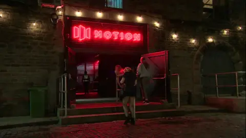 Three people stand outside the entrance to the nightclub Motion, which is located at a large warehouse. 