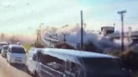A queue of cars can be seen with a big cloud of grey smoke visible behind the powerlines.