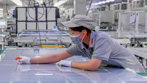 VCG via Getty Images A woman leans over a large sheet of solar panel modules at a factory in Jinhua, Zhejiang Province of China.  
