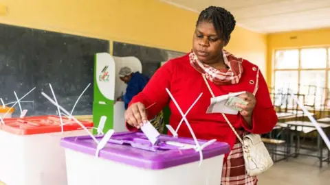 Reuters Inside a school classroom in Lusaka, Zambia, a woman wearing a red top places a ballot paper in a plastic ballot box with a purple lid. Some desks can be seen behind her. 
