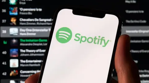 Getty Images A photo of a hand holding a phone which has the green Spotify logo on it. Behind is a Spotify playlist in the background.