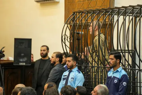 NurPhoto via GettyImages Ateb Najib stands inside a cage wearing a striped uniform as he is sentenced in a courtroom in Damascus, Syria.