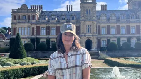 Lord Somerleyton Lara Somerleyton stands in front of a fountain outside her home, Somerleyton Hall, a brick and cream stone Victorian-era mansion.
Lawns, pruned shrubbery and hedges are next to the fountain. A path and steps lead from the fountain to the entrance. 