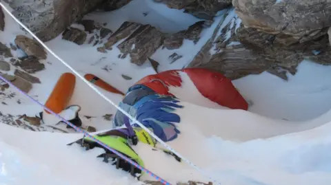 Supplied The dead body of a climber lying face down and partly covered by snow. His green boots can be seen on his feet.
