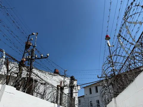 Blue sky above white walls covered in barbed and razor wire. 