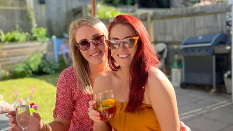 Leanne and Sarah/BBC Friends Sarah (left) and Leanne (right) wearing sunglasses, smiling and holding drinks as they sit in a garden.