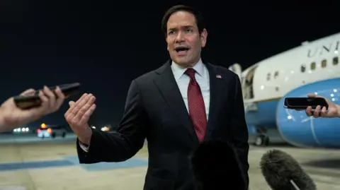 A man gestures with his right hand wearing a dark suit in front of an American government plane, microphones enter the frame.