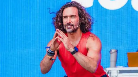 Camp Bestival Joe Wicks, a man with long and dark curly hair, strikes a pose on stage, crouching and placing his hands together. He is wearing a red tank top. 