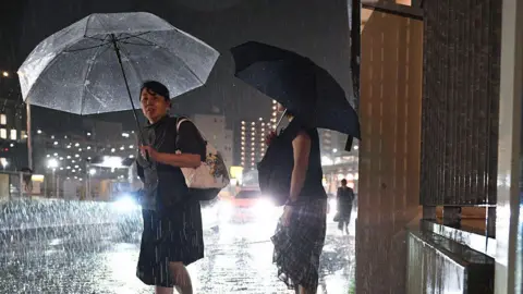 AFP via Getty Images Two women with umbrellas waiting in the rain at night