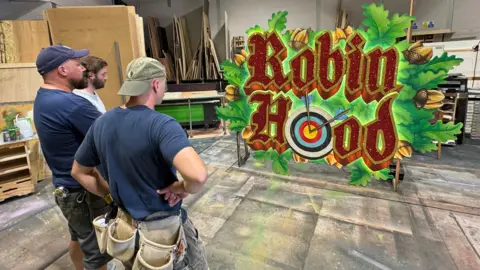 BBC Three men wearing work clothes and navy T-shirts stand with their backs to the camera looking at a large sign reading "Robin Hood". One of the O's is a target with arrows in, and there are oak leaves around the words.