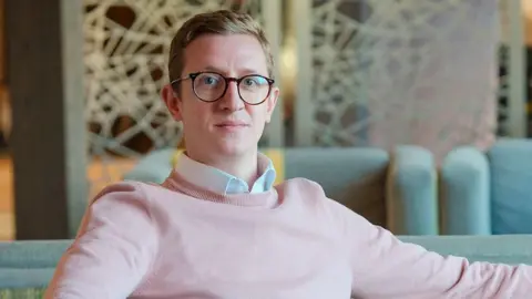Alex West A man wearing glasses and a light pink jumper while seated on a grey sofa in the foreground. One arm rests along the back of the sofa and the other on the armrest. Additional sofas and a patterned room divider are visible in the background of the indoor lounge area.