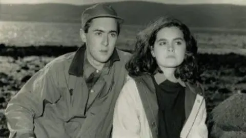 Supplied A black-and-white photograph shows Allan and Shirley seated among large rocks on a shoreline, with water and distant hills visible in the background.
Both are dressed in outdoor clothing, creating a candid, natural scene in a rugged coastal setting.
