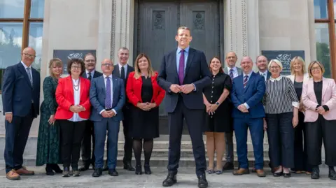 The first minister and cabinet ministers stand on steps in front of a grey building with a brown door.