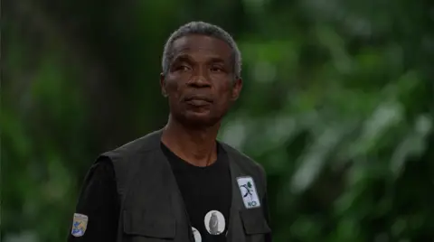 Ifiokabasi Ettang / BBC Chukwuemeka Igwe, stands in the sacred forest in Awka-Etiti. He wears an NCF badge on his gilet and a T-Shirt with photos of vultures.