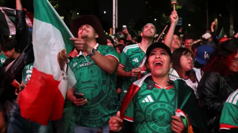 Fans wear Mexico jerseys as they smile and celebrate, some are holding the country's flag.