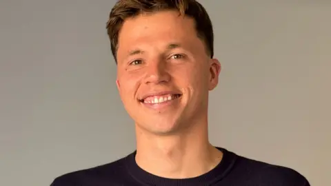 BBC Freddie Woodward: a young man with short dark hair sits on a grey sofa, smiling at the camera. He has short brown hair and wears a navy jumper.