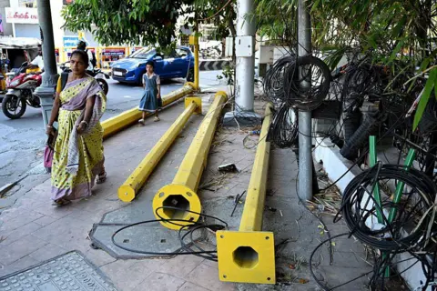 AFP via Getty Images A woman walks past poles and wires lying on a pedestrian footpath in Chennai 