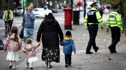 Getty Images A family including two young girls in white dresses and pink coats along with a little boy in a blue coat. They walk down the street with their backs to the camera and in the background two Metropolitan Police officers in uniform can be seen walking in the same direction.