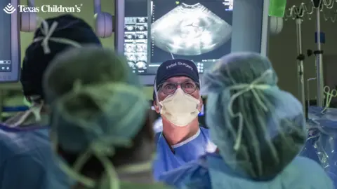 Texas Children’s Hospital Medical staff in the foreground wearing surgical gowns, face masks and hair covers, gathered around an operating table. A person at the centre wears glasses, a face mask and a surgical cap labelled “Fetal Surgery”. Behind the team, large medical monitors display ultrasound images and diagnostic data. IV poles with hanging fluid bags and surgical instruments are visible at the sides of the operating theatre. The text “Texas Children’s” appears in the top-left corner of the image.