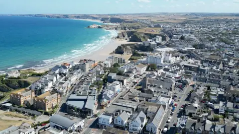 BBC An aerial drone shot of Newquay town centre in the sunshine, with blue skies and the beach on the left, a very blue sea with white waves - and cliffs and countryside stretching into the distance. The buildings are largely grey and white and there are criss-crossing roads visible with cars on them.