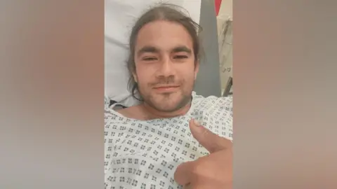 Paul Byrne A man in hospital gown and bed, he has long dark hair
