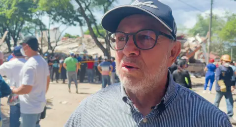 BBC News Mundo A man with a short white beard, glasses, a baseball cap, and a buttoned up shirt with an oxford collar 