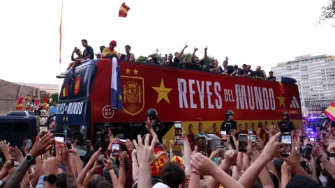 An open-top bus carries the Spain national football team through the streets of Madrid. Crowds cheer in celebration from the street.