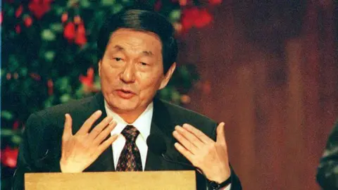 Boston Herald via Getty Images Then-Chinese Premier Zhu Rongji, wearing a suit and sitting behind a desk, giving a speech at the Massachusetts Institute of Technology in the US in 1999