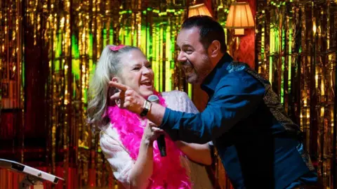 BBC/Jack Barnes/Kieron McCarron A blonde woman smiles broadly while wearing a pink feather boa and holding a karaoke microphone. She is stood on a glittery stage with a brown hair man who is also smiling. 