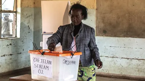 AFP via Getty Images A woman in a patterned jacket and African wrap for a skirt stands at a ballot box  putting her vote in. She is in an empty classroom and a blackboard can be seen behind her.