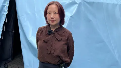 Sachie Mikawa A Japanese woman stands in front of a light blue fabric backdrop with hands behind their back. She is wearing a brown long-sleeved blouse with a bow-tie neckline and dark trousers