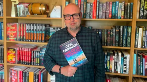 BBC A man standing in front of giant book case full of books, he is wearing a checked shirt and black glasses. He is holding a book in his hands