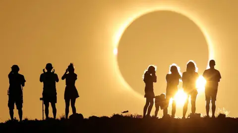 Reuters A total solar eclipse is about to occur - a darkened grey sphere appears large in the sky and bright yellow light is emanating from it drenching the sky. Two groups are gathered watching it, silhouetted against the sky 