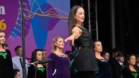 Pacemaker A woman wearing black clothing stands with an arm outstretched. She has long dark hair, and there are more people behind her, also wearing dark clothing. 