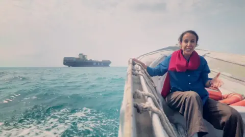 Nawal wears a scarf, shirt and trousers as she sits in a boat on the Strait with a shipping container seen in the background