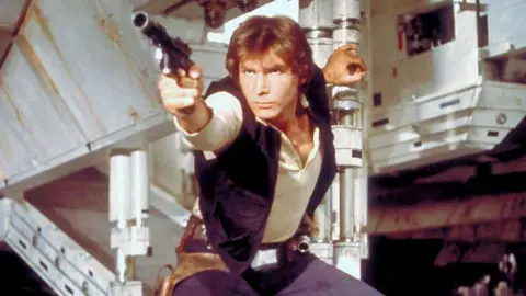 Getty Images Han Solo in a screen grab from Star Wars. He has a phaser in hand about to shoot and is holding the open exit ramp to the millennium falcon 