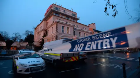 CAROLINE QUINN/GETTY IMAGES A garda (Irish police) cordon outside the Regency Hotel in Dublin in 2016.  A white and blue liveried garda van and car are parked outside a four-storey hotel at dusk on a wet evening.  A white a blue cordon tape stretches across an entrance to the hotel car park. 