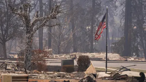 Reuters A burned United States flag hangs from a charred flagpole - burned trees and remnants of destroyed properties can be seen on either sides of the road and the air is hazy 